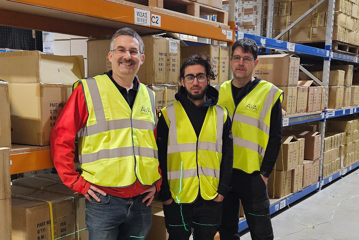 Hussain Asif pictured in a manufacturing facility, with shelves of boxes behind him. He is between two other men.